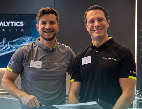 Two WAA Consultants smiling and standing behind a desk, wearing matching branded polo shirts. The man on the left wears a gray shirt, and the man on the right wears a black shirt with yellow accents.